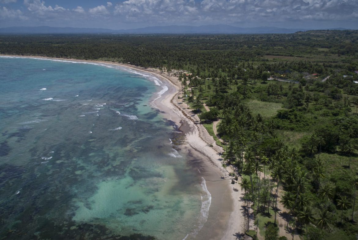 buyDRproperty Aerial view of a tropical beach with turquoise water and palm trees, showcasing oceanfront development property potential. Sosua Real Estate
