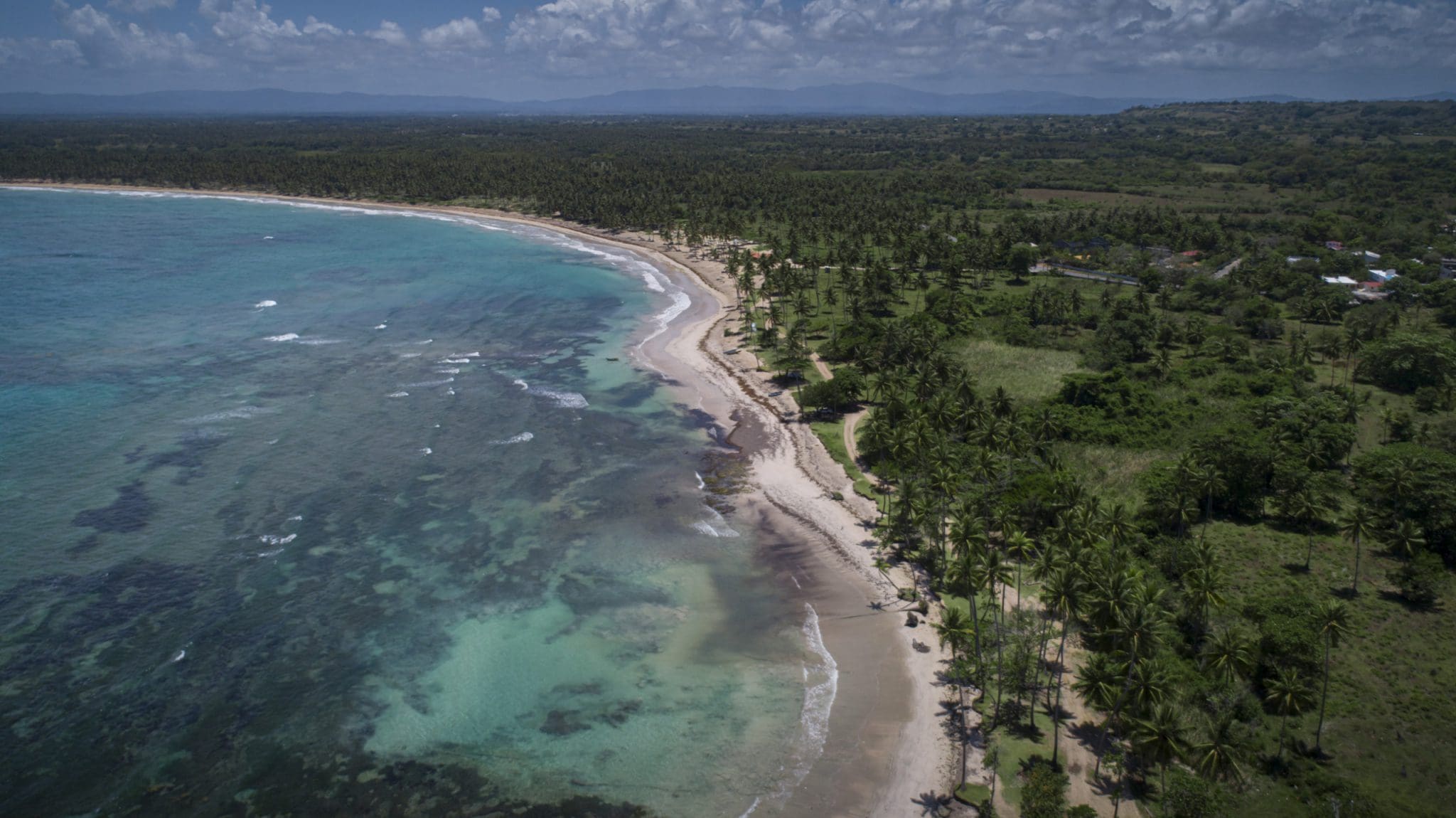 buyDRproperty Aerial view of a tropical beach with turquoise water and palm trees, showcasing oceanfront development property potential. Sosua Real Estate