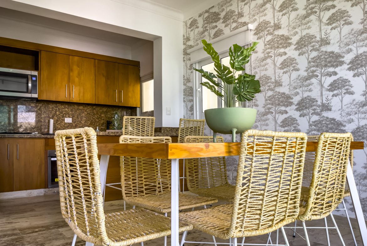 buyDRproperty Modern dining area with wooden table and woven chairs. Kitchen in background. Sosua Real Estate