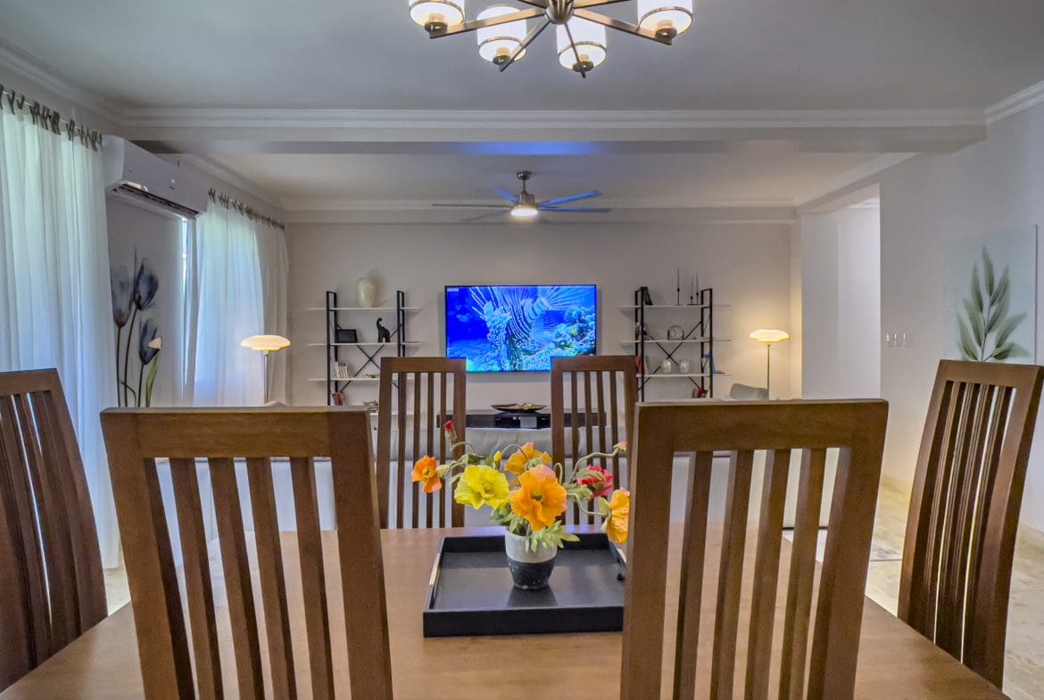 buyDRproperty Dining room of a 3 Bedroom Oceanfront Condo with wooden table and chairs, flowers, and a TV. Sosua Real Estate