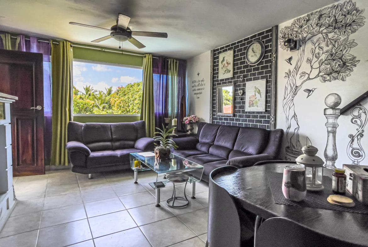 buyDRproperty Living room of a 2-bedroom condo in Sosua with dark purple couches, glass table, and tree mural. Sosua Real Estate