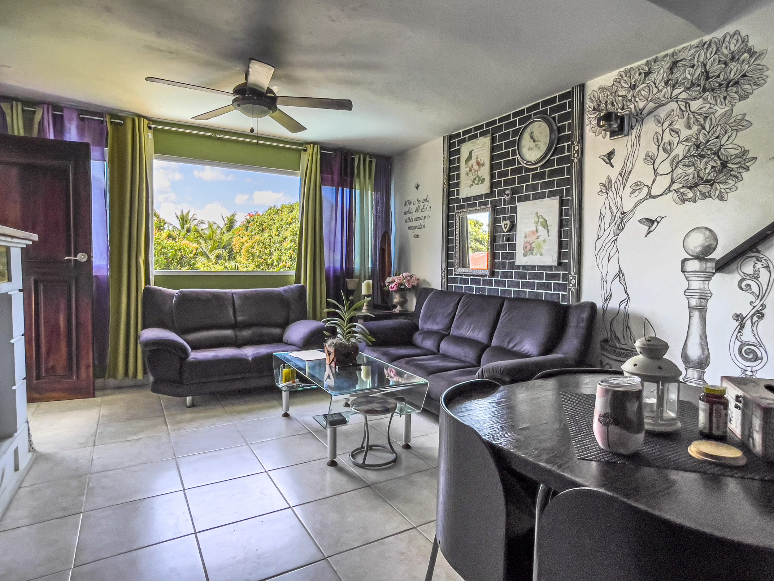 buyDRproperty Living room of a 2-bedroom condo in Sosua with dark purple couches, glass table, and tree mural. Sosua Real Estate