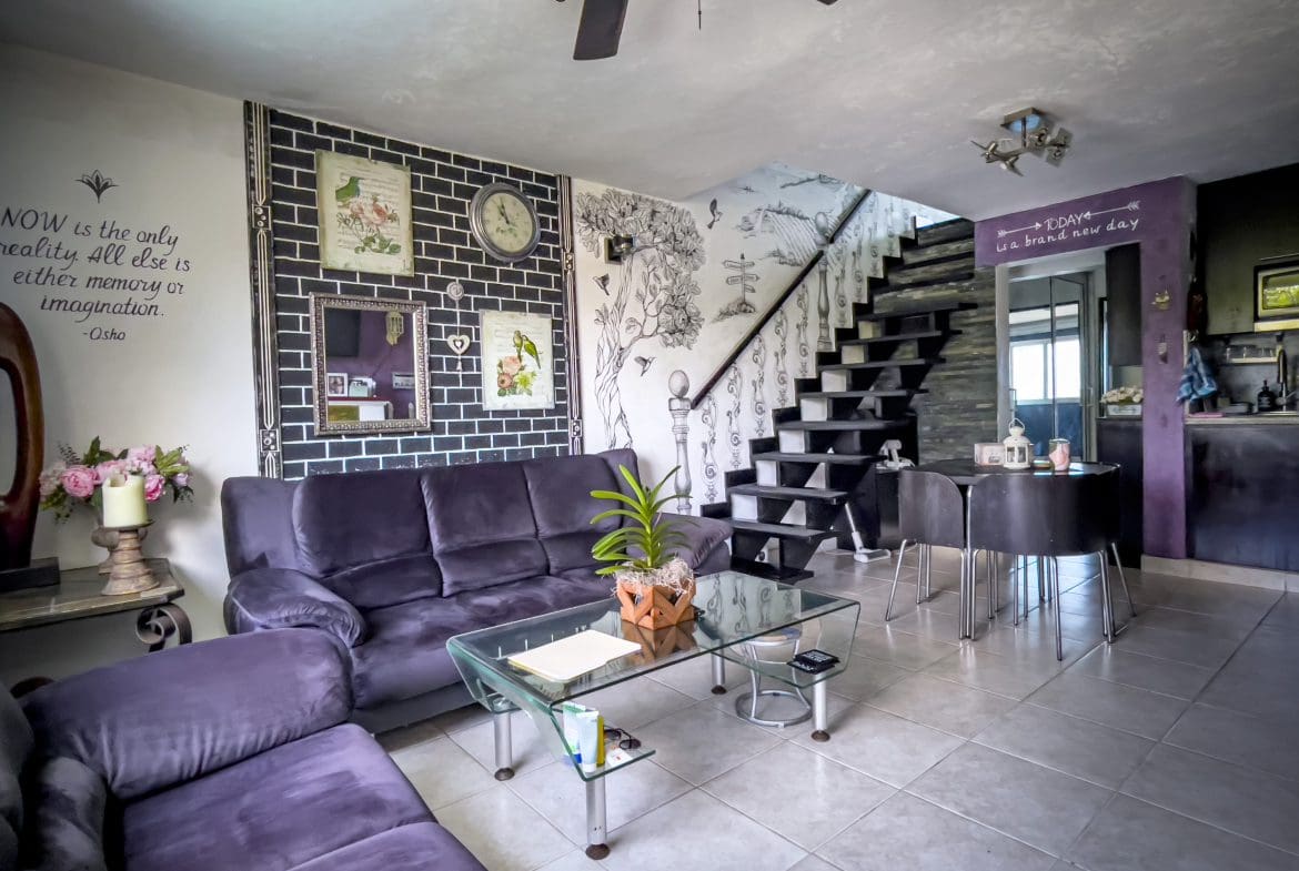 buyDRproperty Purple living room with black brick wall, sofa, and glass coffee table. "Now is the only reality" quote. Sosua Real Estate