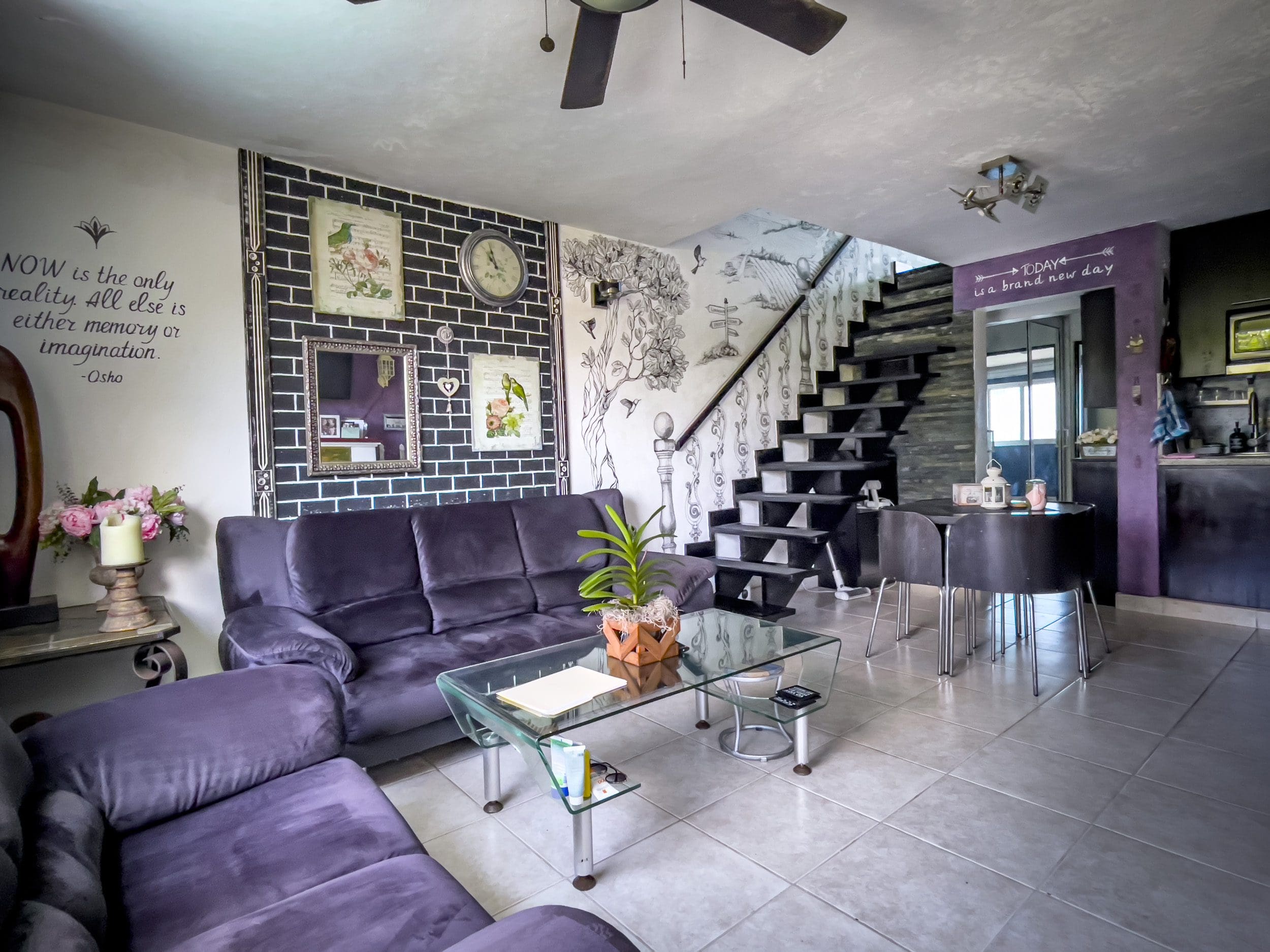 buyDRproperty Purple living room with black brick wall, sofa, and glass coffee table. "Now is the only reality" quote. Sosua Real Estate