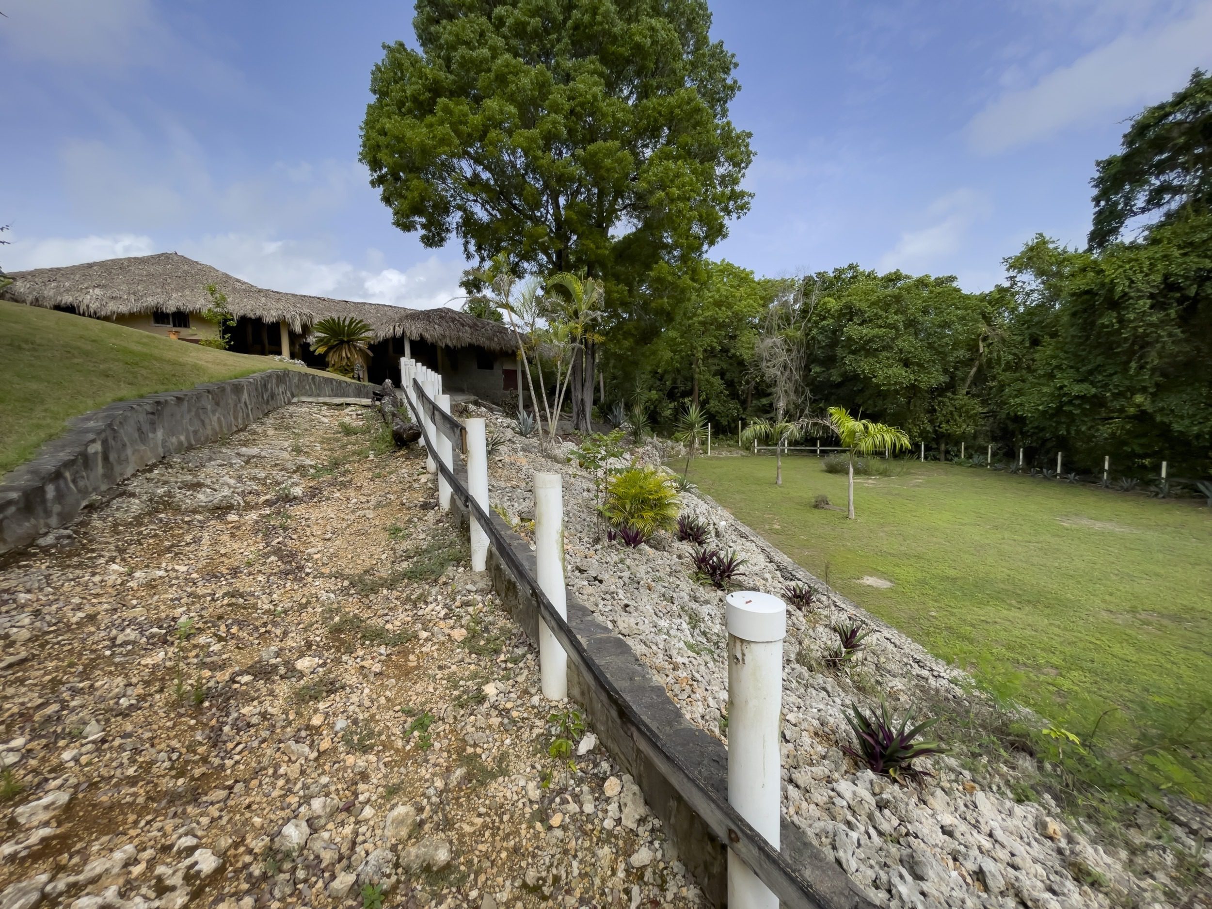 buyDRproperty Pathway leading to a tropical resort with thatched roofs, surrounded by lush greenery and blue sky. Sosua Real Estate