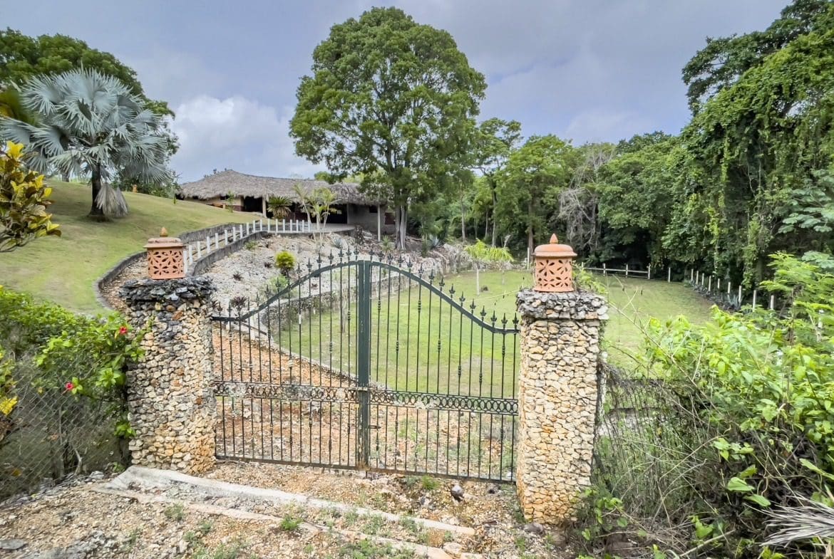 buyDRproperty Gated entrance to building lot with stone pillars, lush landscaping, and thatched roof building. Sosua Real Estate