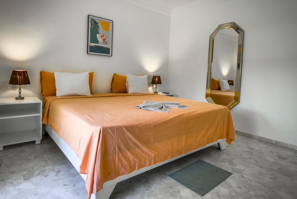 buyDRproperty Modern bedroom with orange bedspread, white pillows, and large mirror; prime villa. Sosua Real Estate