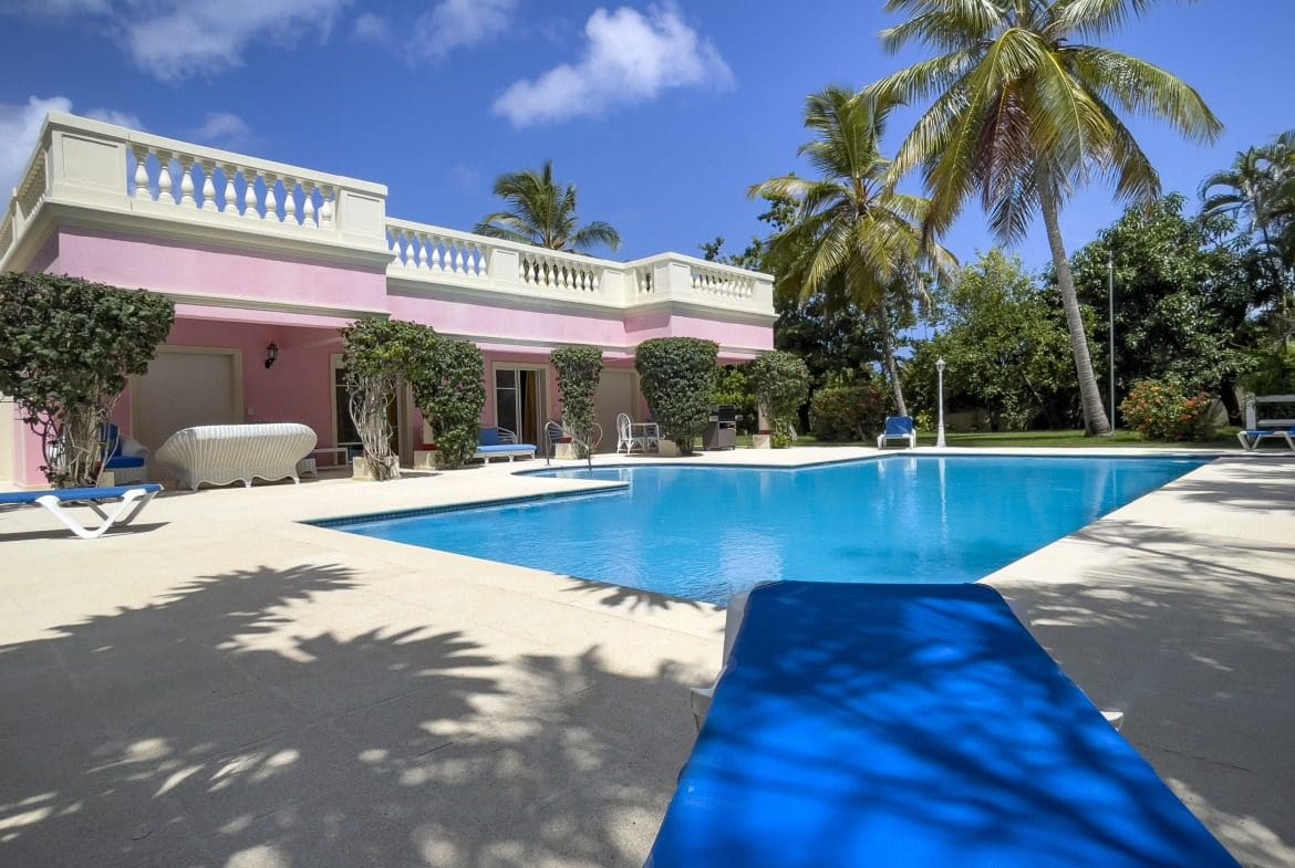 buyDRproperty Prime pink villa with a pool and palm trees under a blue sky. Sosua Real Estate