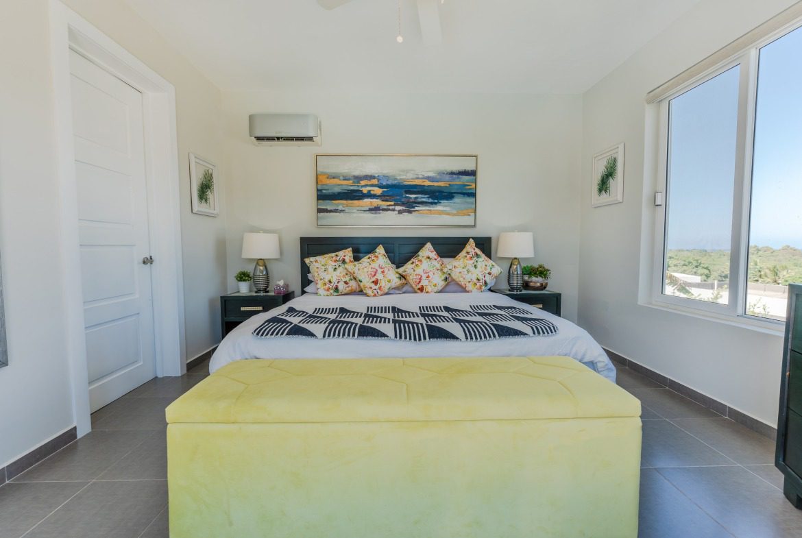 buyDRproperty Master bedroom with a yellow ottoman, artwork, and window overlooking the ocean. Bedroom design and interior design. Sosua Real Estate