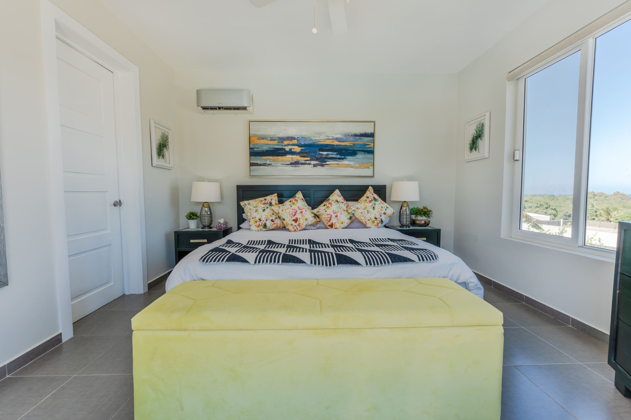 buyDRproperty Master bedroom with a yellow ottoman, artwork, and window overlooking the ocean. Bedroom design and interior design. Sosua Real Estate