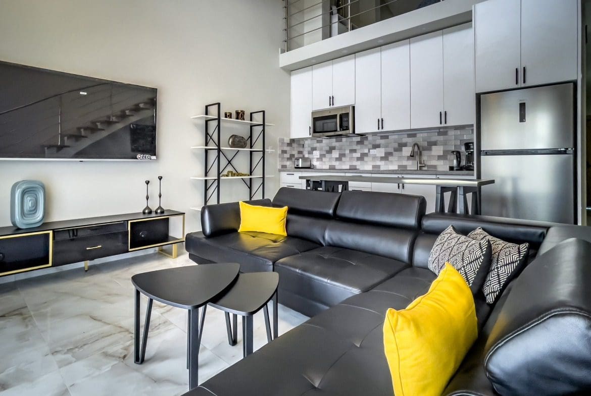 buyDRproperty Modern 1-bedroom condo living room with black leather sectional sofa and kitchen in the background. Sosua Real Estate