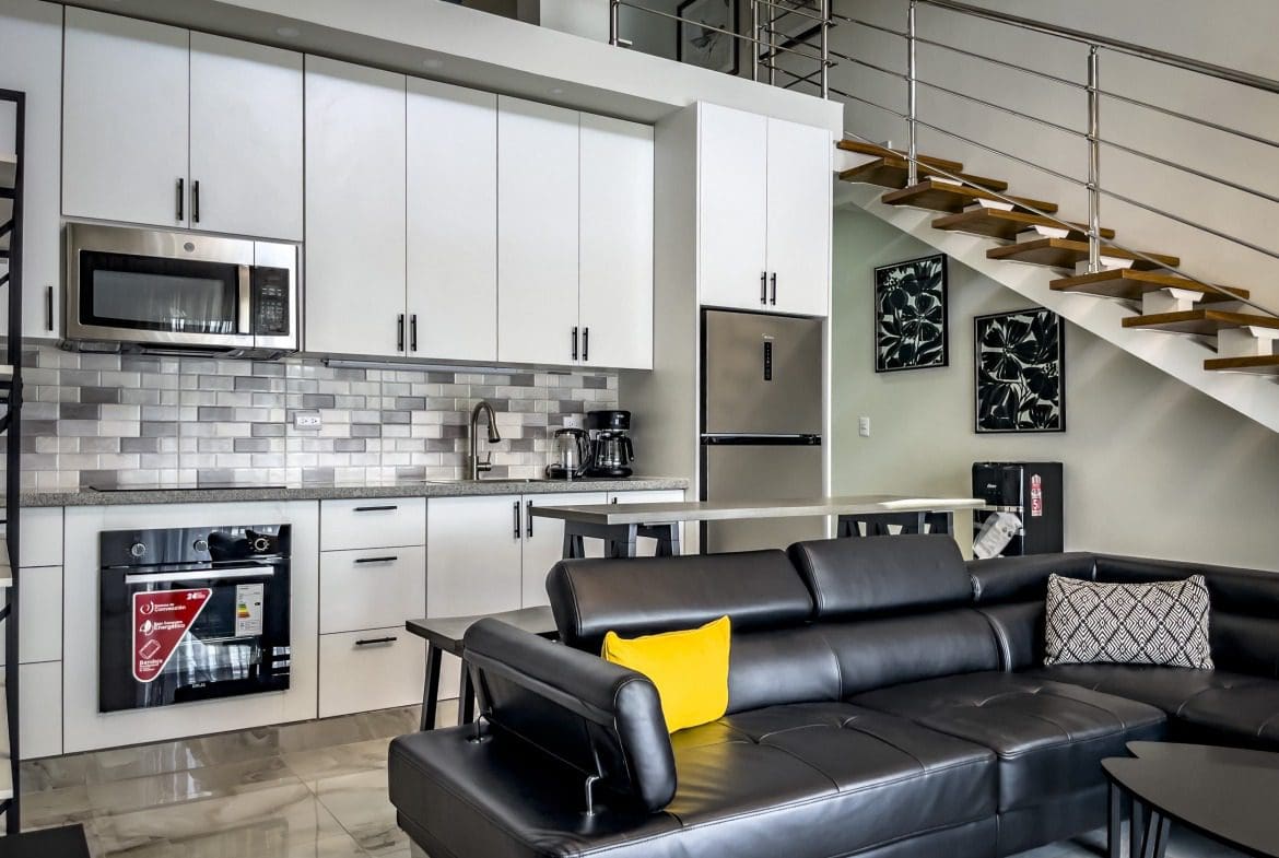 buyDRproperty Modern 1-Bedroom Condo kitchen with white cabinets, stainless steel appliances, and black leather sofa. Sosua Real Estate