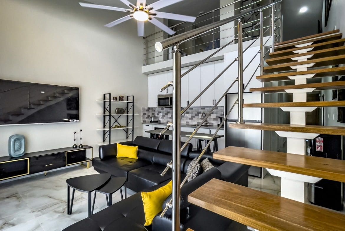 buyDRproperty Stylish 1-Bedroom Condo living room with black leather sofa, loft stairs and modern kitchen. Sosua Real Estate