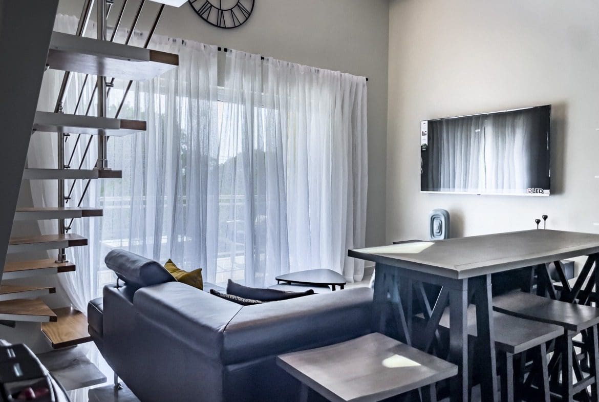 buyDRproperty Modern 1-bedroom condo living room with floating staircase, sofa, and TV. Sosua Real Estate