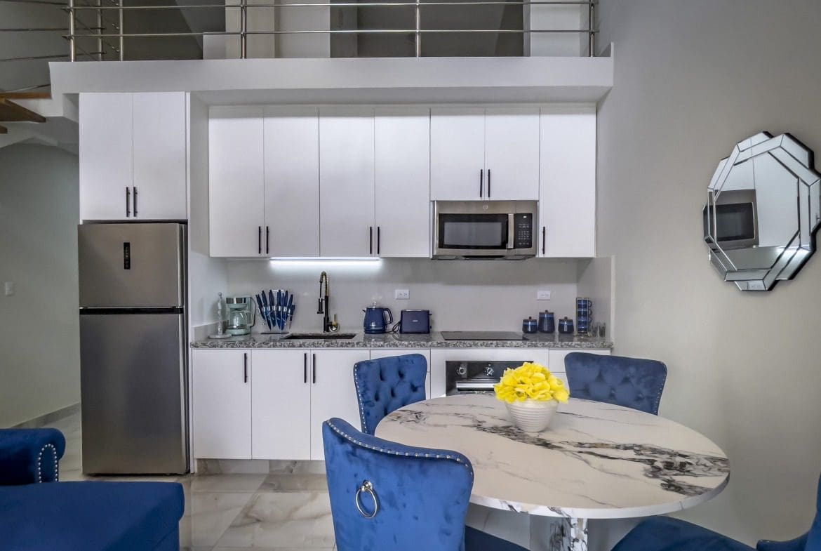 buyDRproperty Modern 1-Bedroom Condo kitchen and dining area featuring white cabinets, stainless steel appliances, and a round table with blue velvet chairs. Sosua Real Estate