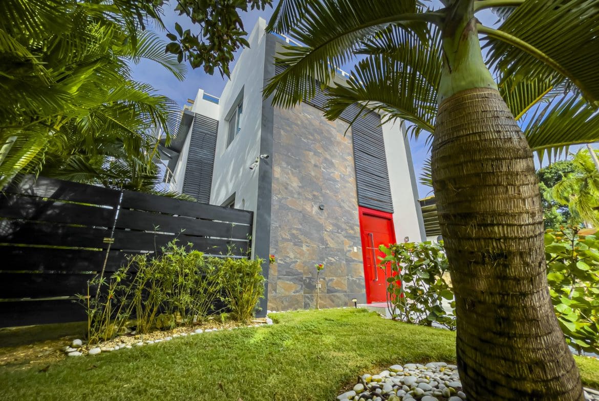 buyDRproperty Modern 3-bedroom villa for sale in Sosúa Ocean Village, Dominican Republic, featuring a red door and lush landscaping. Sosua Real Estate