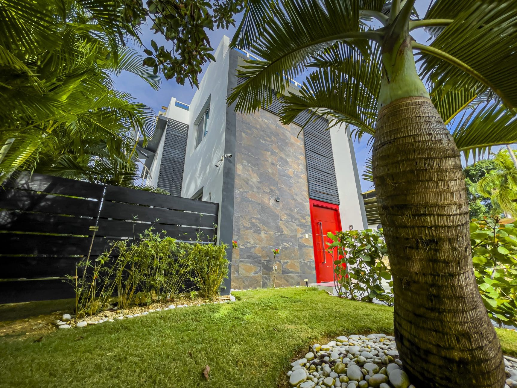 buyDRproperty Modern 3-bedroom villa for sale in Sosúa Ocean Village, Dominican Republic, featuring a red door and lush landscaping. Sosua Real Estate