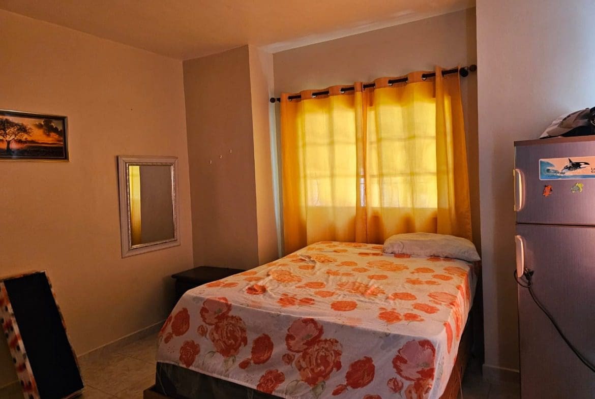 buyDRproperty Bedroom with floral bedspread, yellow curtains, and refrigerator. Sosua Real Estate
