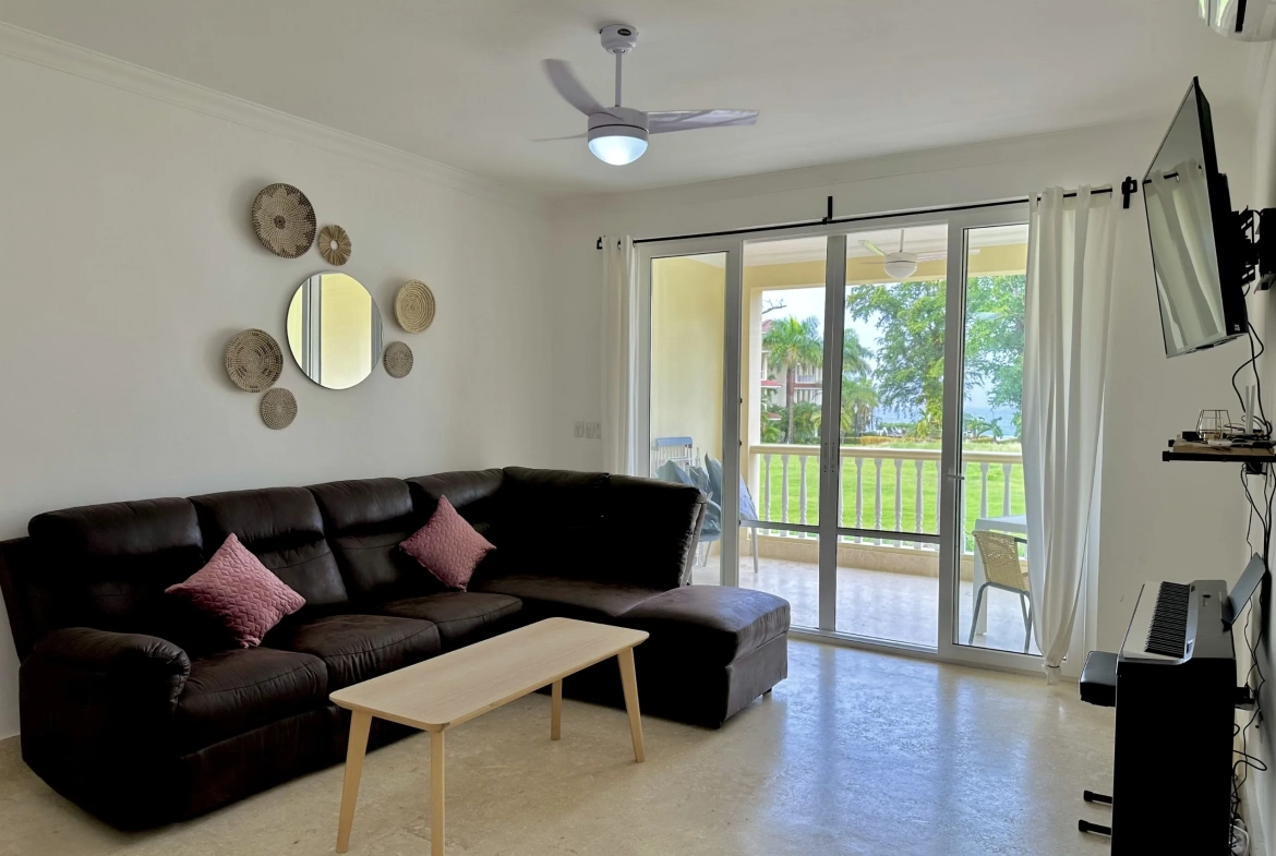 buyDRproperty Living room of a 2-bedroom condo for sale, featuring a dark leather sectional sofa and balcony access. Sosua Real Estate