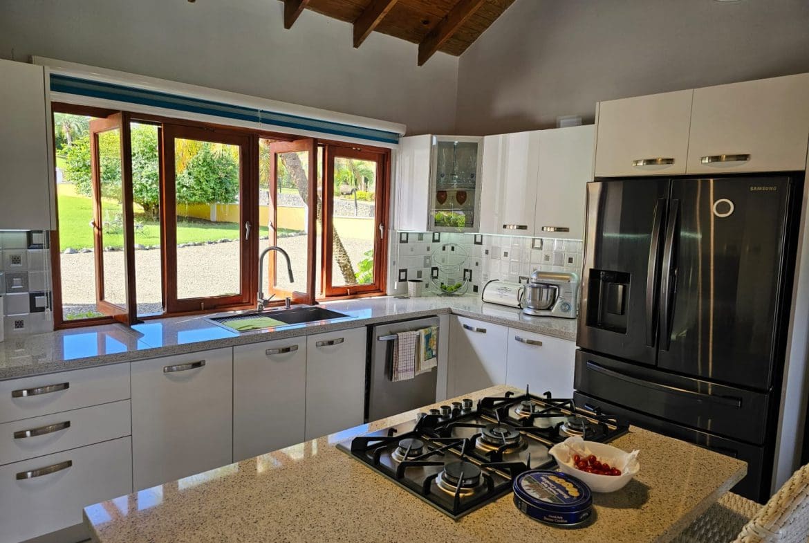buyDRproperty Modern kitchen with island gas cooktop and window showing lush outdoor view of a luxury villa. Sosua Real Estate