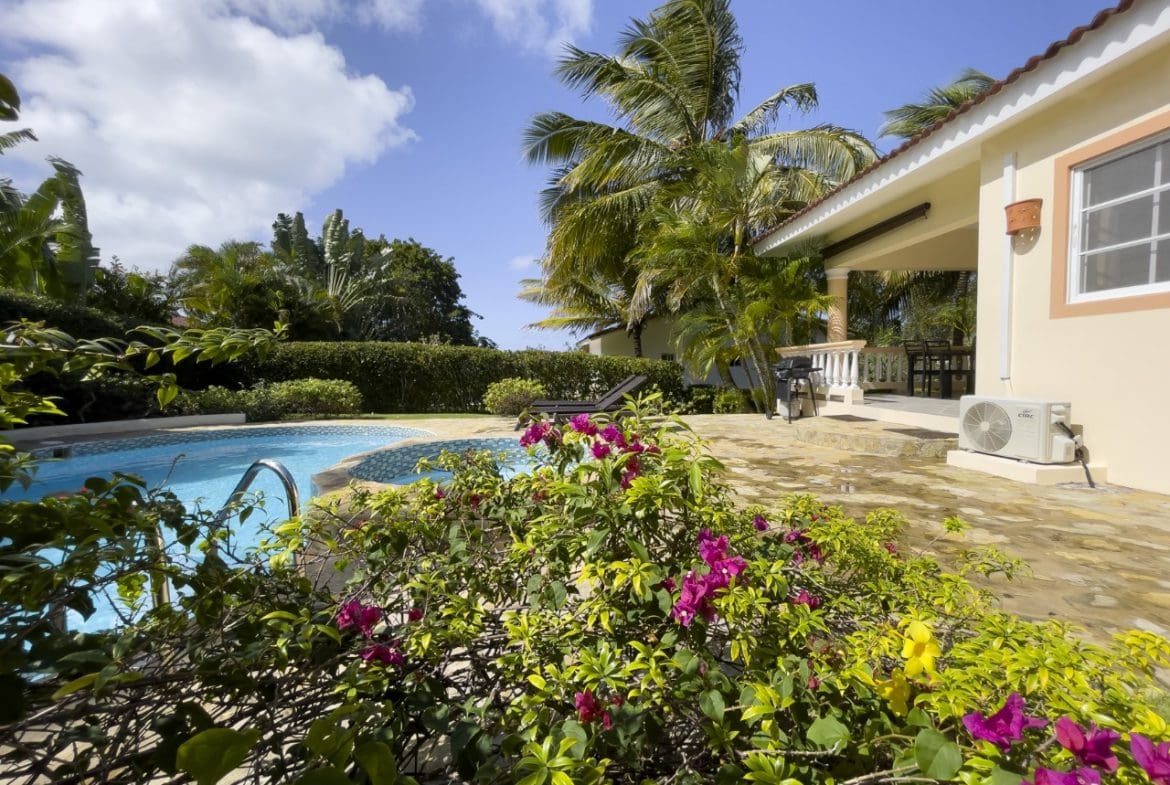 buyDRproperty Poolside view of a 3BR Ocean View Home in Hispaniola, lush tropical garden, and blue sky. Sosua Real Estate