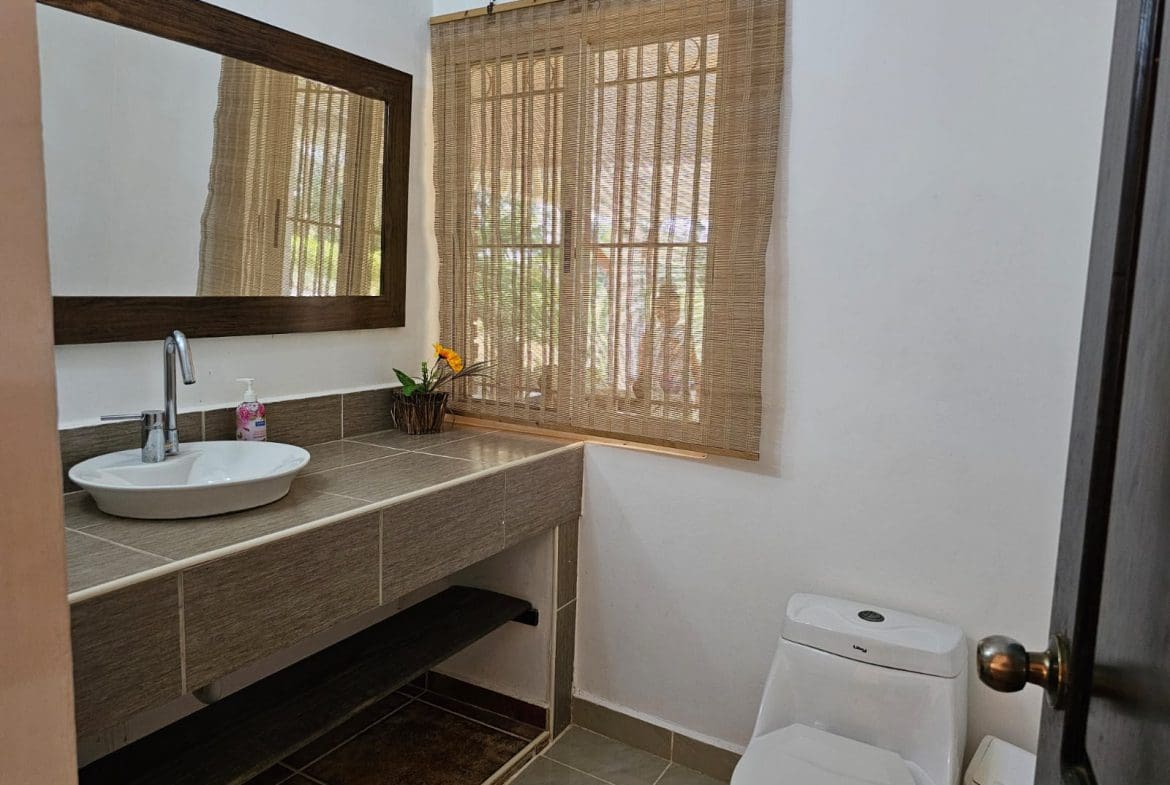 buyDRproperty Bathroom with a modern sink, mirror, woven window shade, and toilet in a Sosua villa. Sosua Real Estate