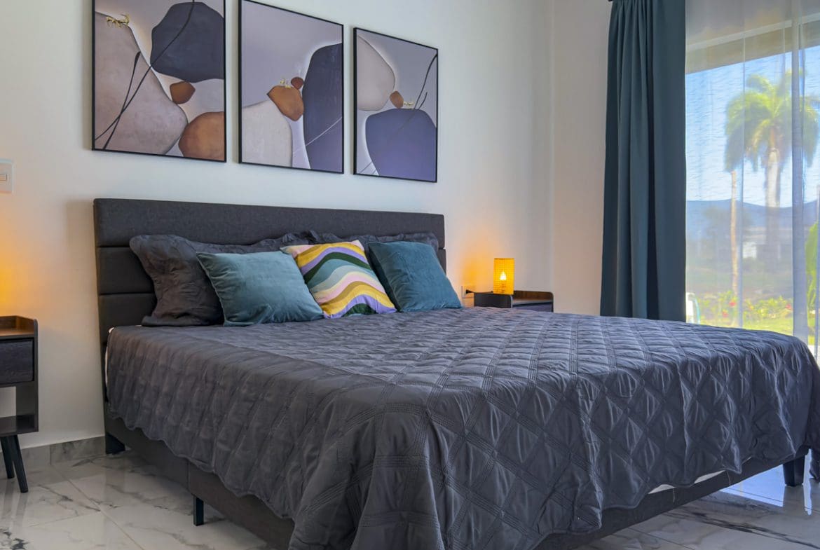 buyDRproperty Modern bedroom with a gray bed, bedside lamps, and artwork. View of palm trees. Sosua Real Estate