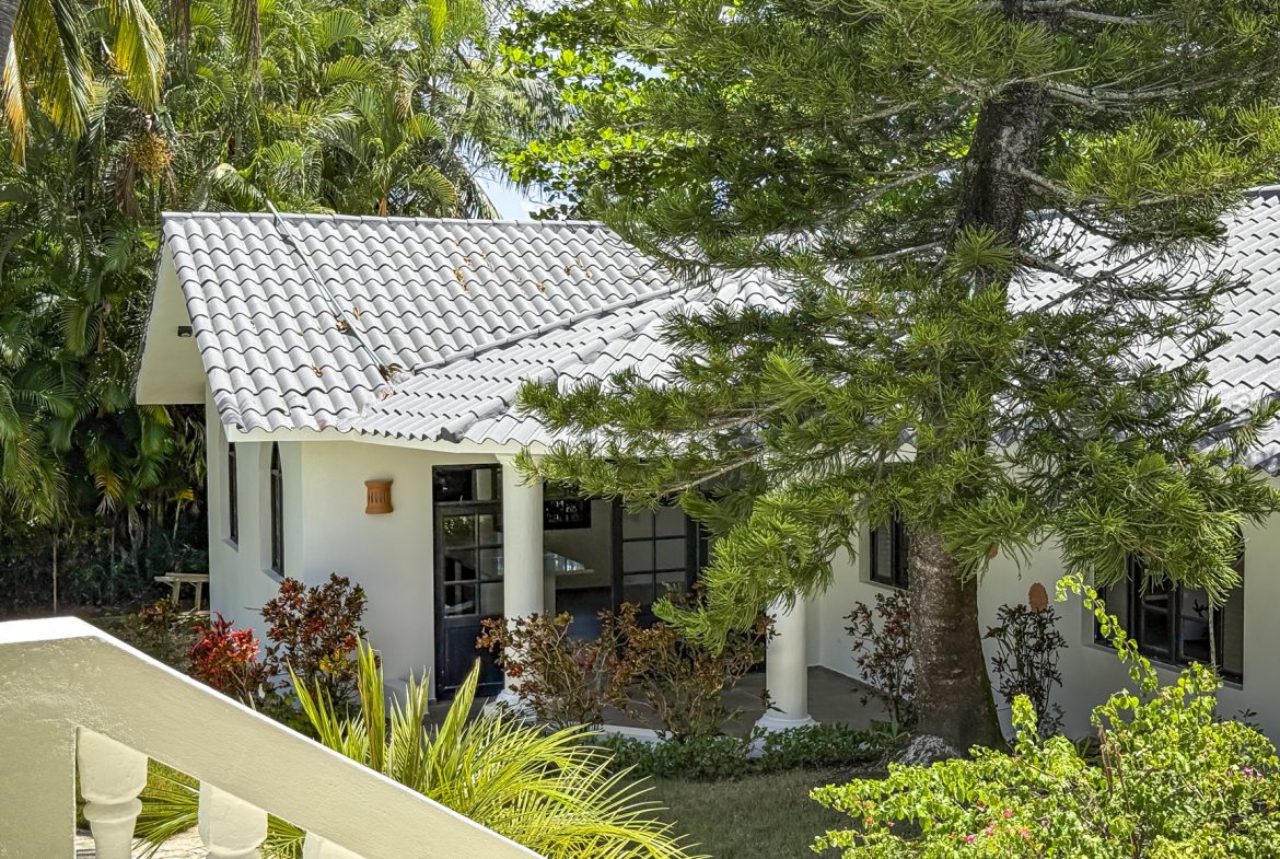 buyDRproperty White stucco vacation home with gray tile roof surrounded by lush tropical greenery. Sosua Real Estate