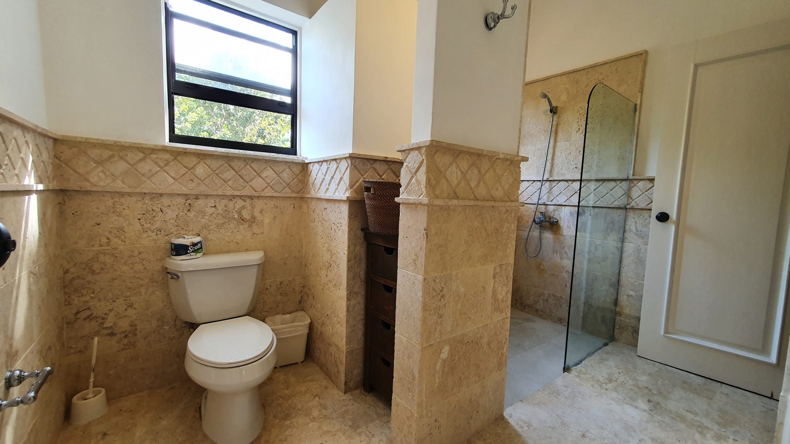 buyDRproperty Travertine tile bathroom with toilet, shower, and natural light from window. Sosua Real Estate