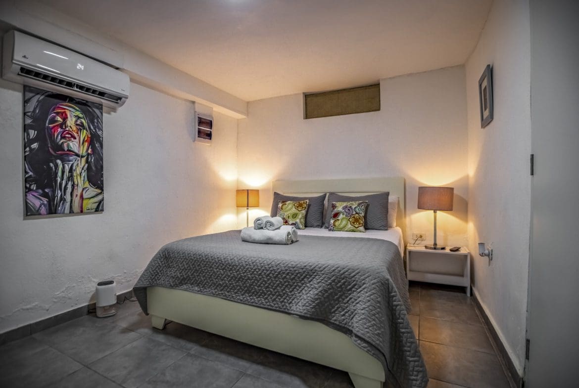 buyDRproperty Cozy bedroom with queen bed, gray quilt, bedside lamps, and modern wall art. Sosua Real Estate