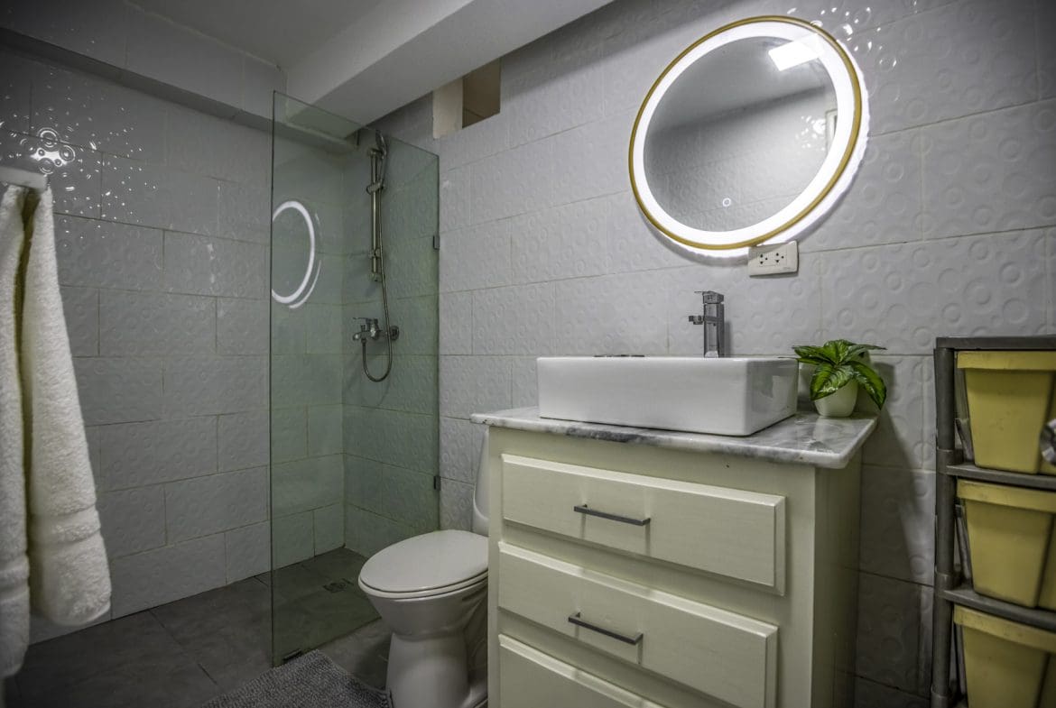 buyDRproperty Modern bathroom with white square tiles, round mirror, vessel sink, and glass shower enclosure. Sosua Real Estate