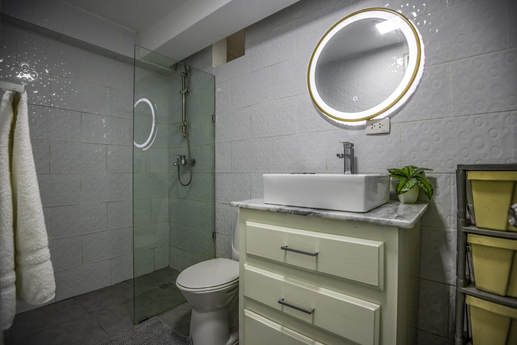 buyDRproperty Modern bathroom with white square tiles, round mirror, vessel sink, and glass shower enclosure. Sosua Real Estate