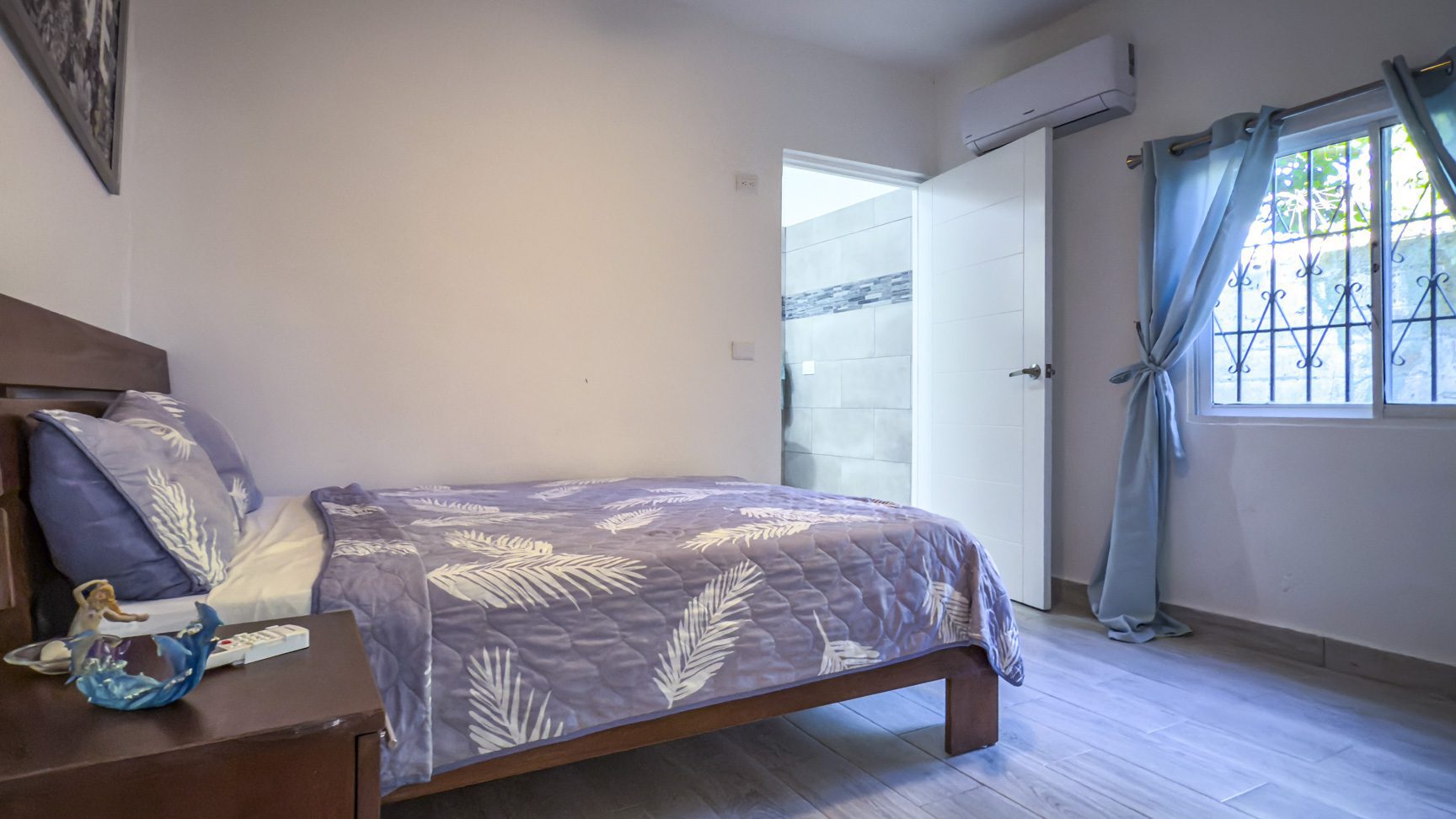 buyDRproperty Cozy bedroom with double bed, blue bedding, and bathroom access. Natural light from window. Sosua Real Estate