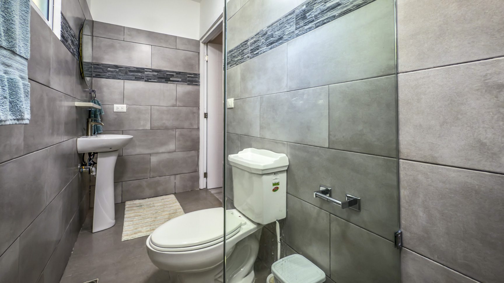 buyDRproperty Modern bathroom with gray tile, pedestal sink, toilet, and glass shower enclosure. Sosua Real Estate