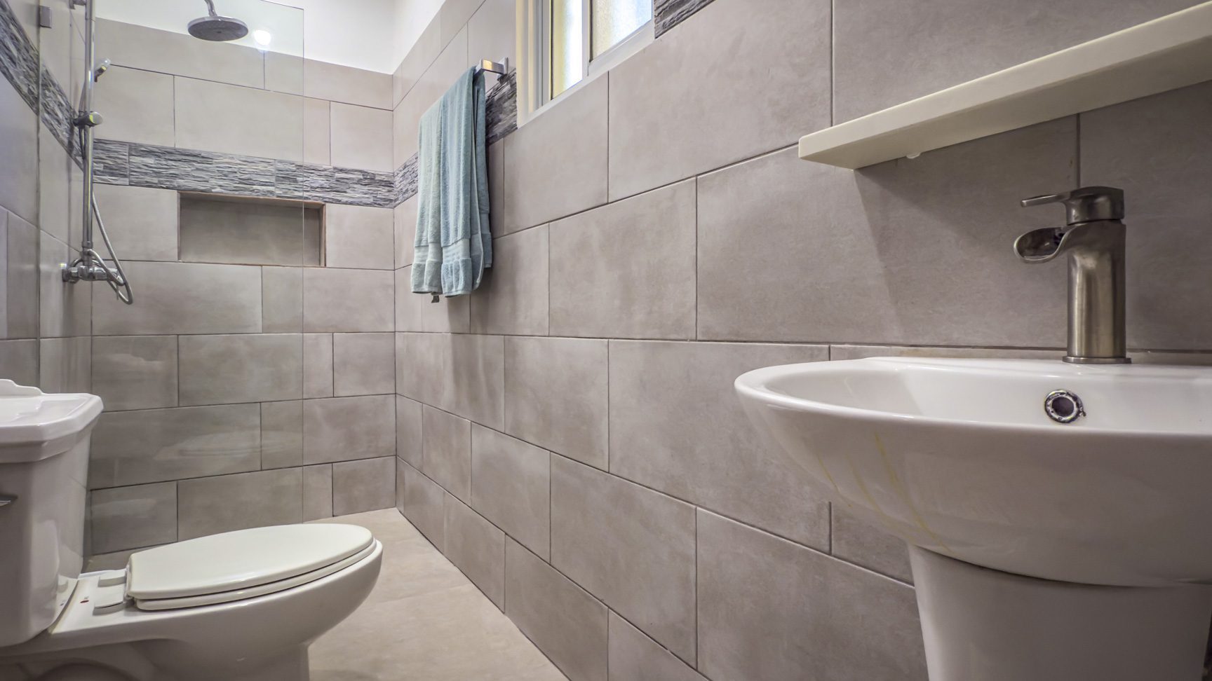 buyDRproperty Modern bathroom with gray tile, white toilet, and pedestal sink under a window Sosua Real Estate