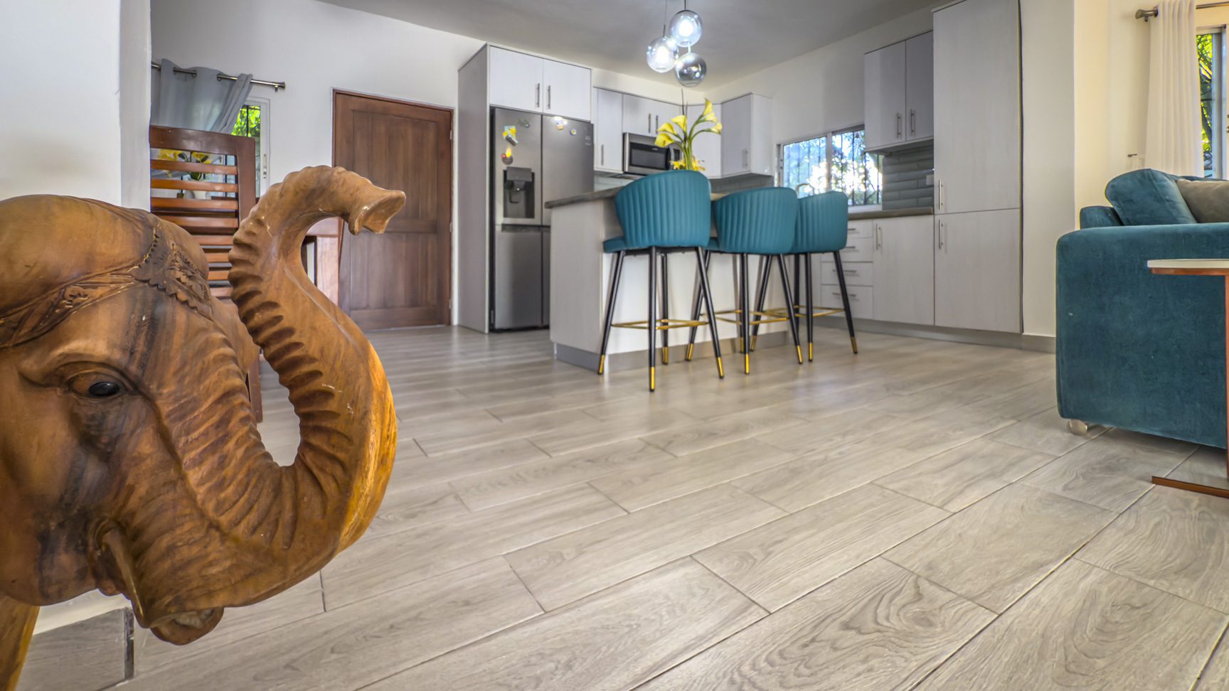 buyDRproperty Bright, modern kitchen with teal bar stools, light wood floors, stainless steel appliances, and a wooden elephant sculpture. Sosua Real Estate