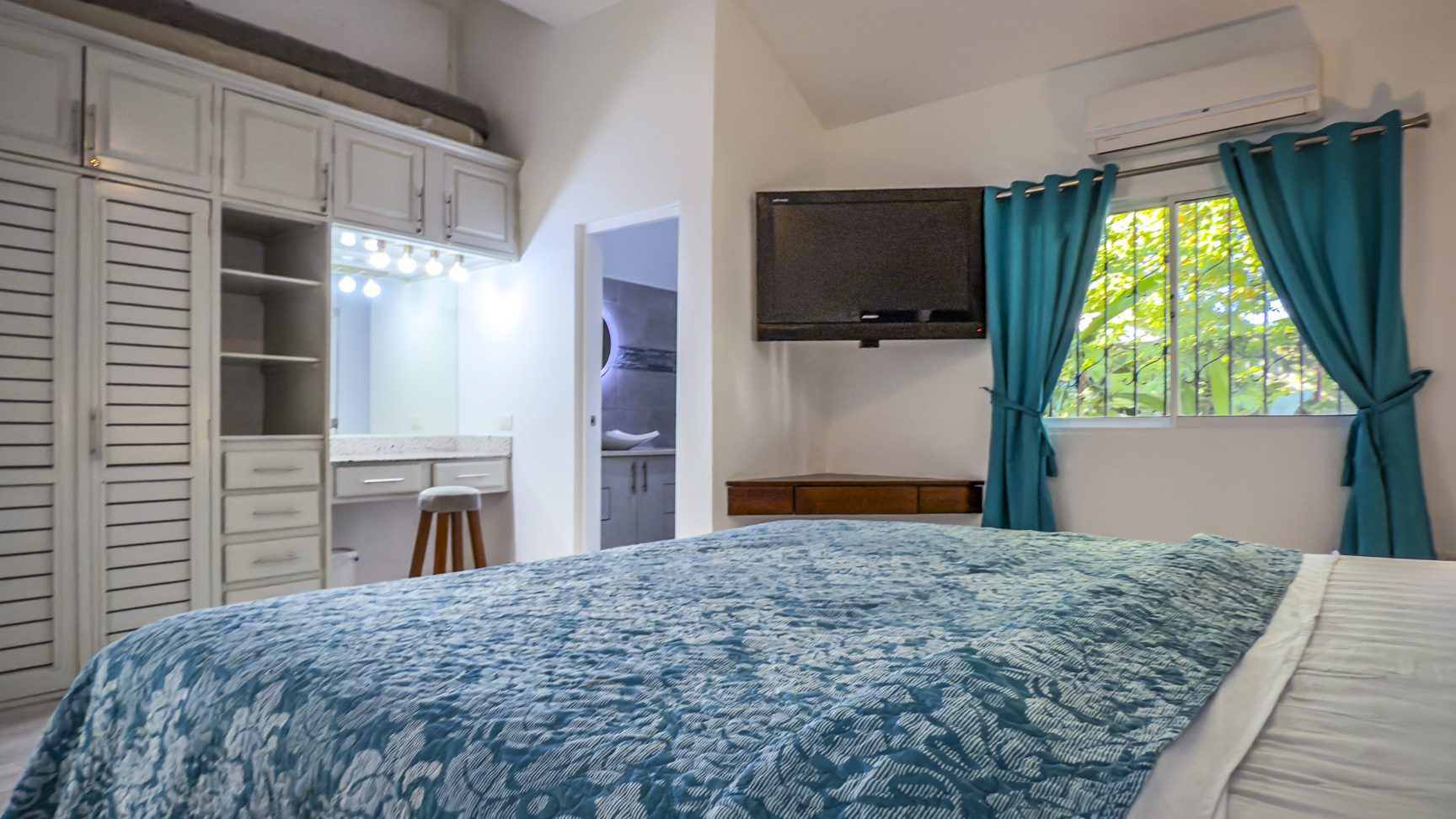 buyDRproperty Bedroom with blue quilt, TV, and window with turquoise curtains. Vanity with lights and stool. Sosua Real Estate