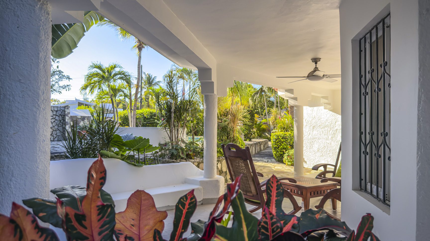 buyDRproperty Covered patio with white columns, tropical plants, and outdoor furniture, offering a relaxing view of the garden. Sosua Real Estate