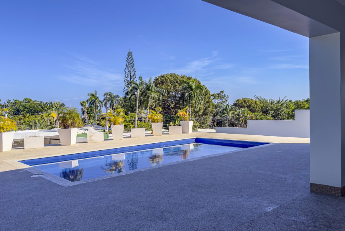 buyDRproperty La Mulata villa pool: Rectangular pool with potted plants and lush tropical trees under a clear blue sky. Sosua Real Estate