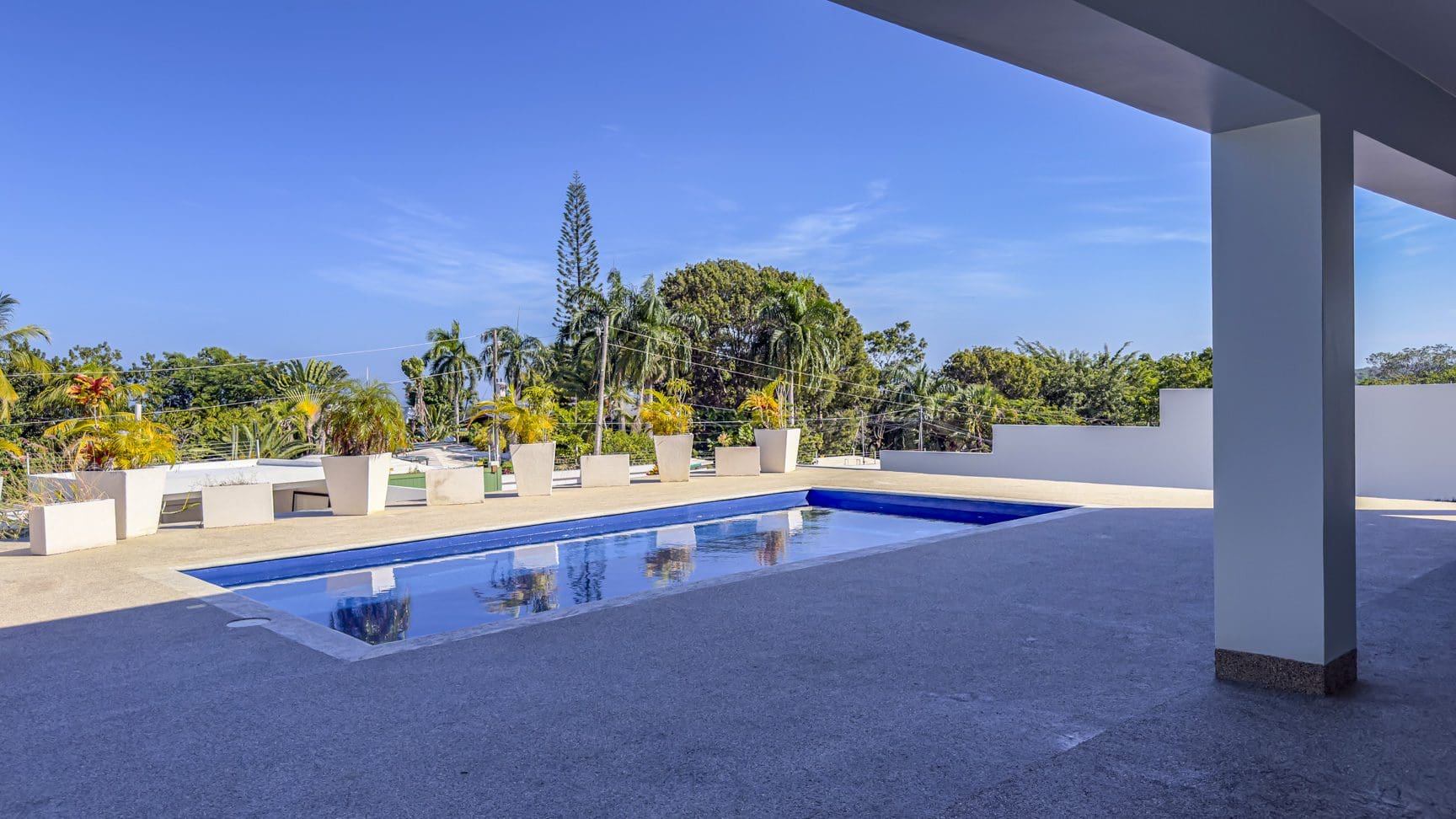 buyDRproperty La Mulata villa pool: Rectangular pool with potted plants and lush tropical trees under a clear blue sky. Sosua Real Estate