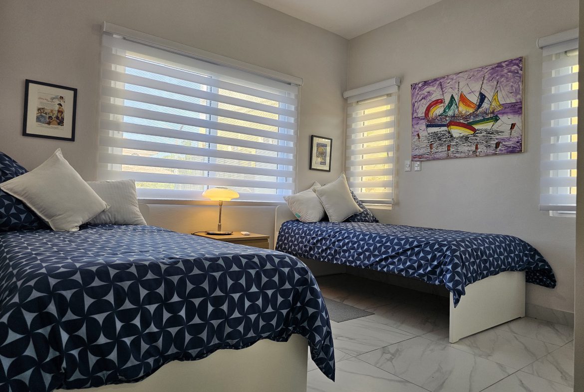 buyDRproperty Bright bedroom with two twin beds, blue patterned bedding, colorful sailboat painting, and white tile floor. Sosua Real Estate