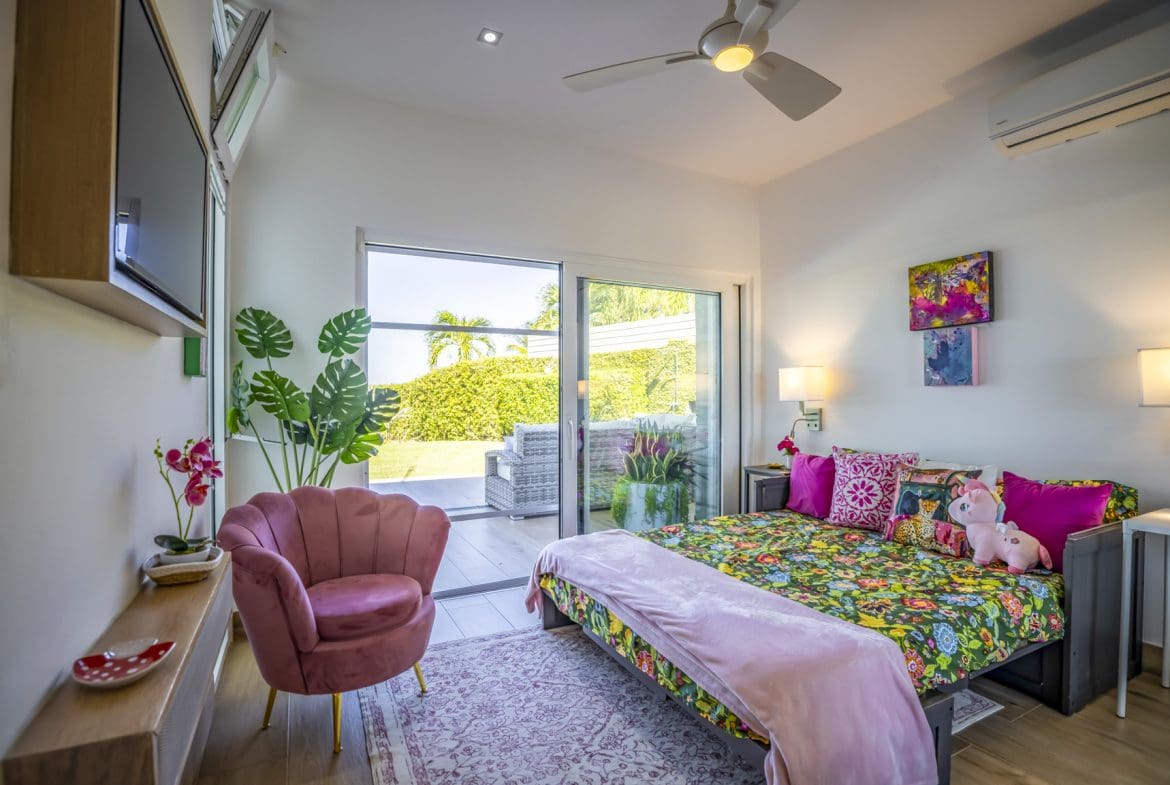 buyDRproperty Bright bedroom with floral bedding, pink accents, open patio doors to garden view, and a pink velvet chair. Sosua Real Estate