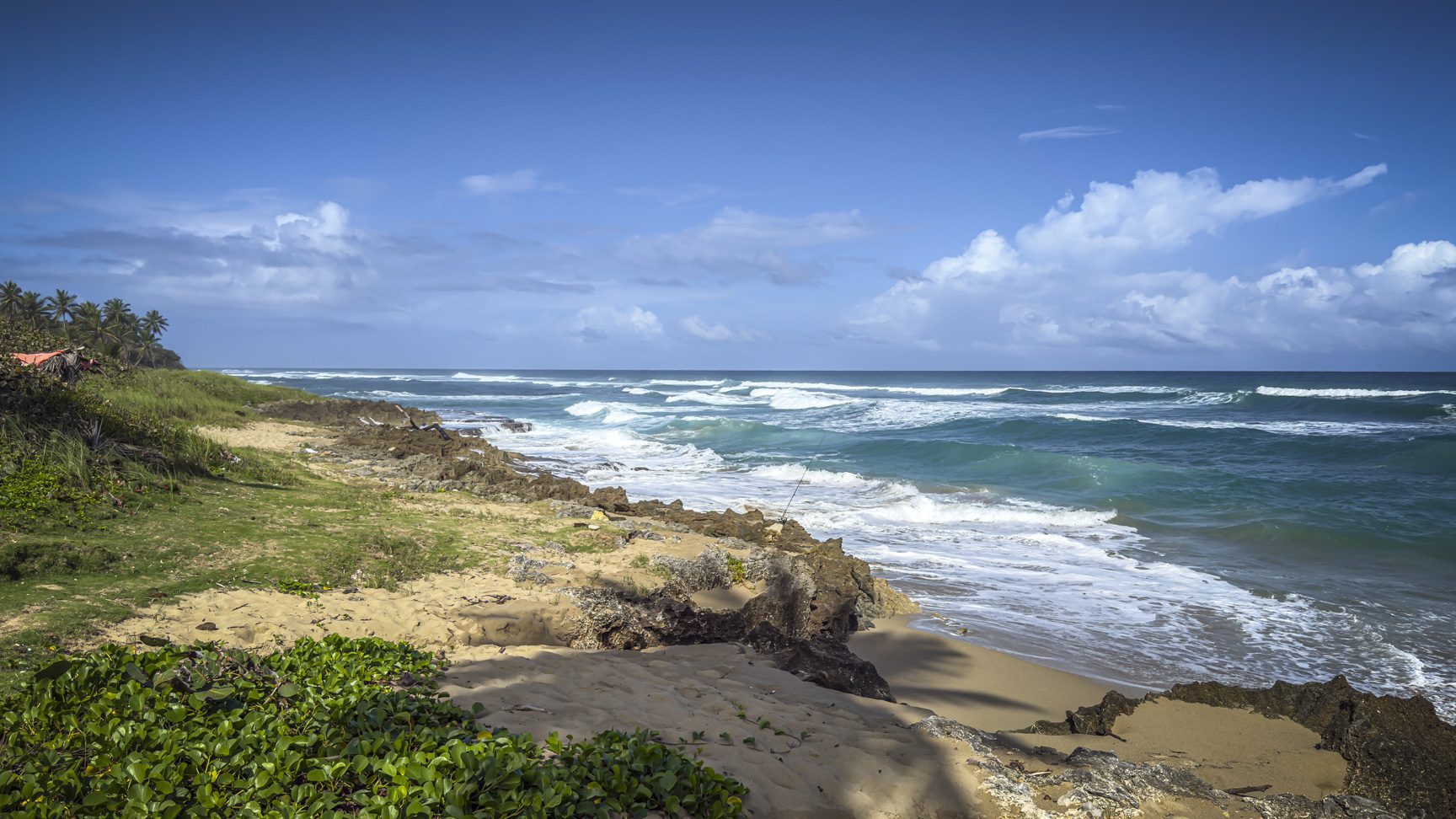 buyDRproperty Turquoise waves crashing on a sandy beach with rocky outcrops and lush green vegetation under a blue sky. Sosua Real Estate