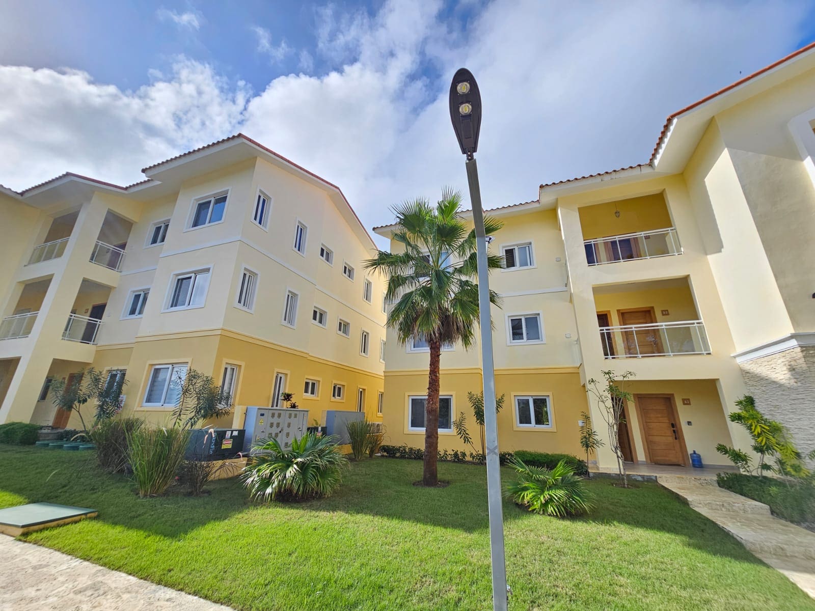 buyDRproperty Modern yellow apartment building with palm tree and green lawn on a sunny day. Sosua Real Estate