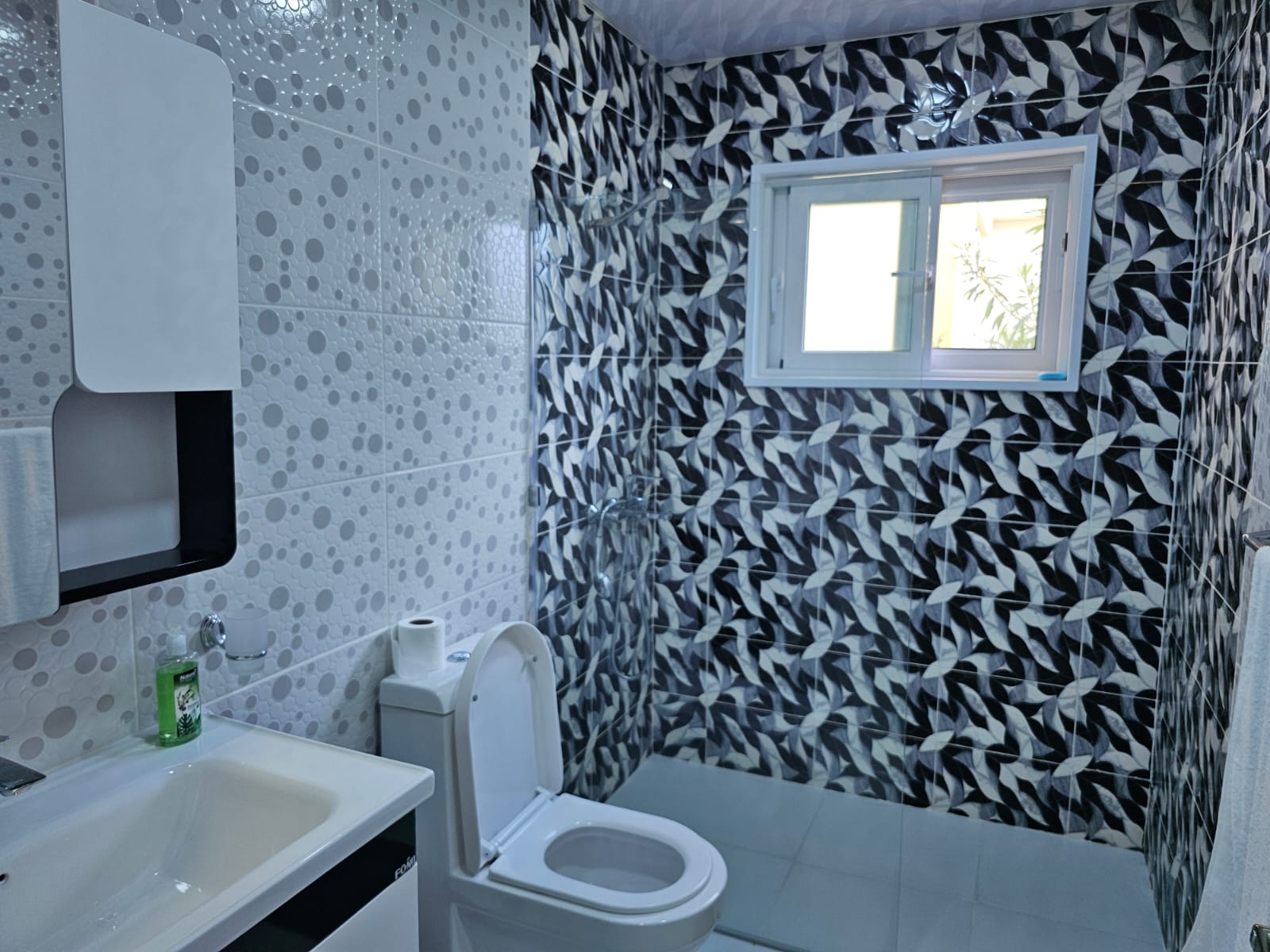 buyDRproperty Modern bathroom with white toilet, sink, and black/white patterned shower tiles, illuminated by natural light. Sosua Real Estate