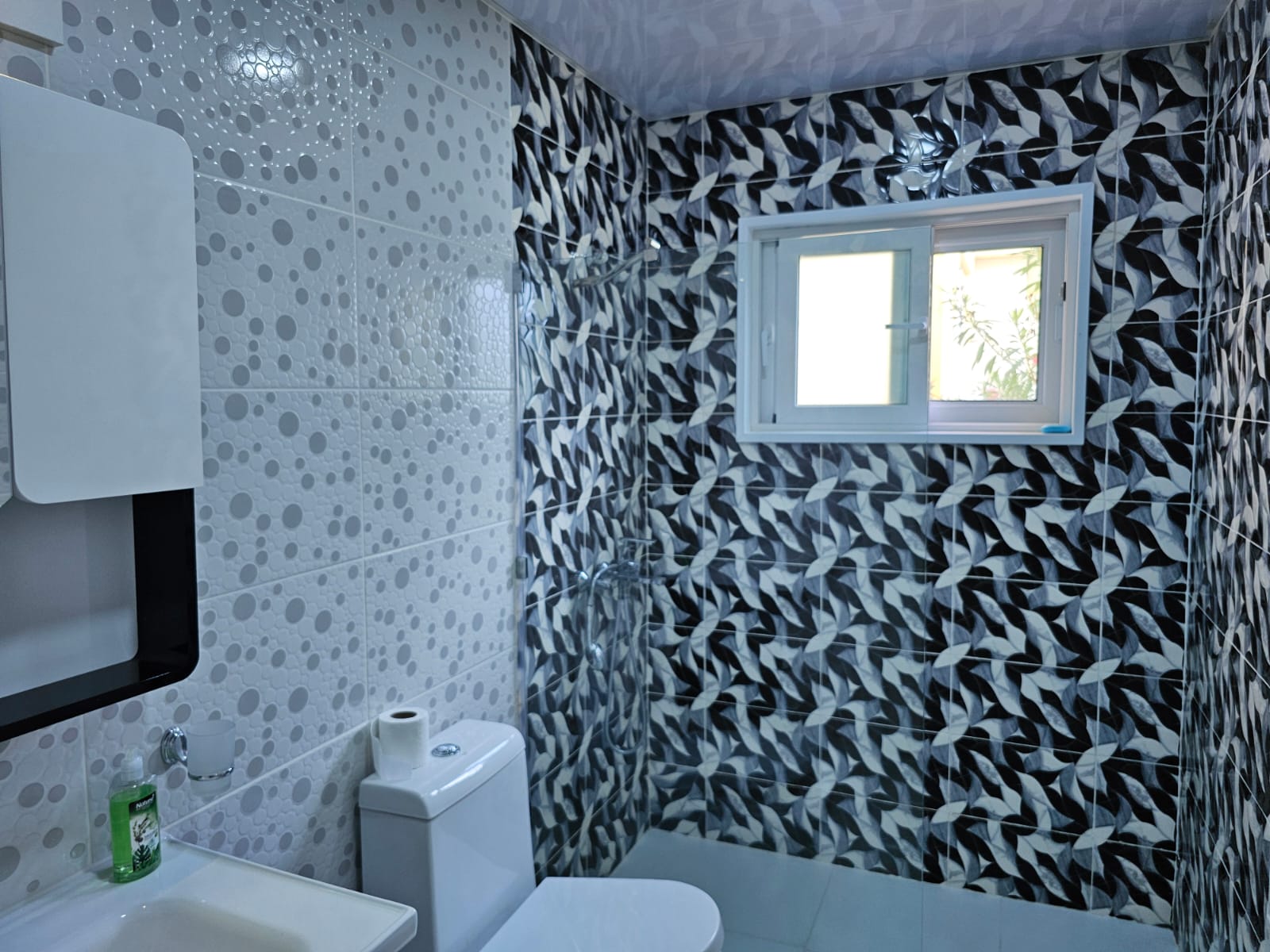 buyDRproperty Modern bathroom with gray patterned tiles, white toilet, sink, and window providing natural light. Sosua Real Estate