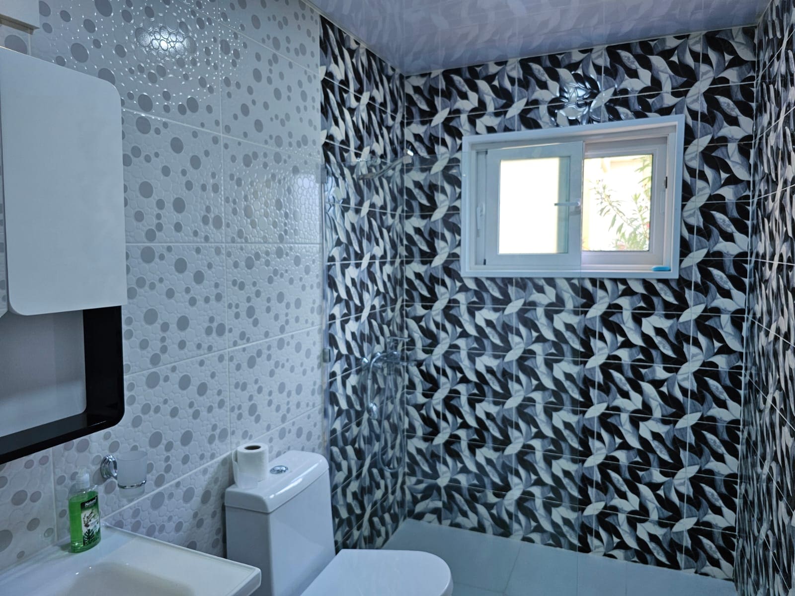 buyDRproperty Modern bathroom with gray patterned tiles, white toilet, sink, and window providing natural light. Sosua Real Estate