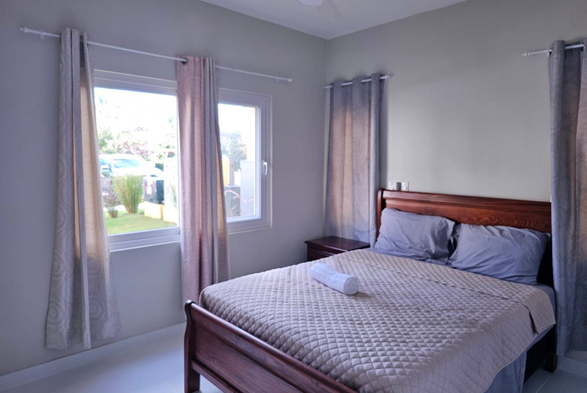 buyDRproperty Cozy bedroom with wooden bed frame, quilted beige bedspread, and two windows with curtains. Sosua Real Estate
