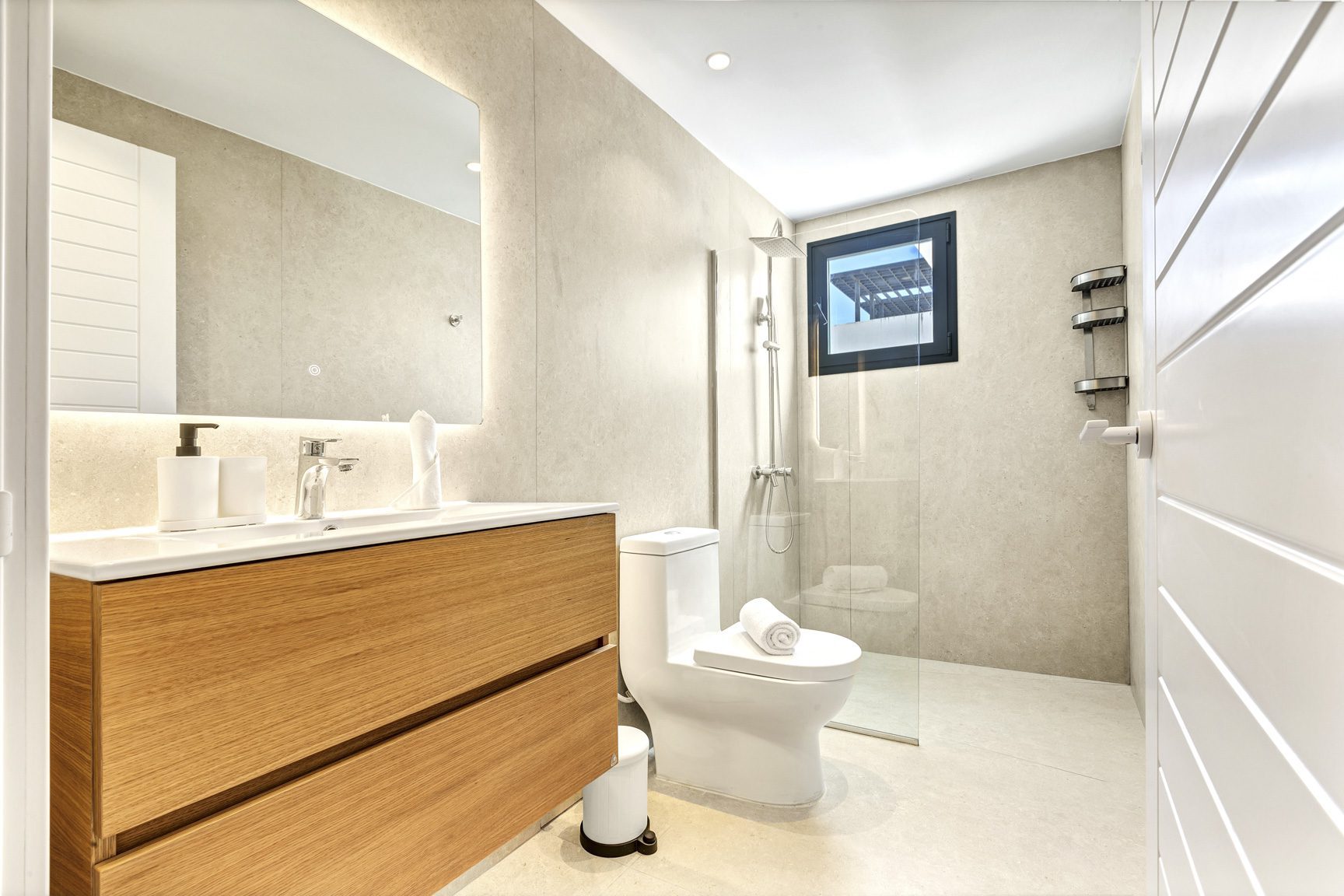 buyDRproperty Modern bathroom with wood vanity, toilet, glass shower, light tile, and white door. Sosua Real Estate