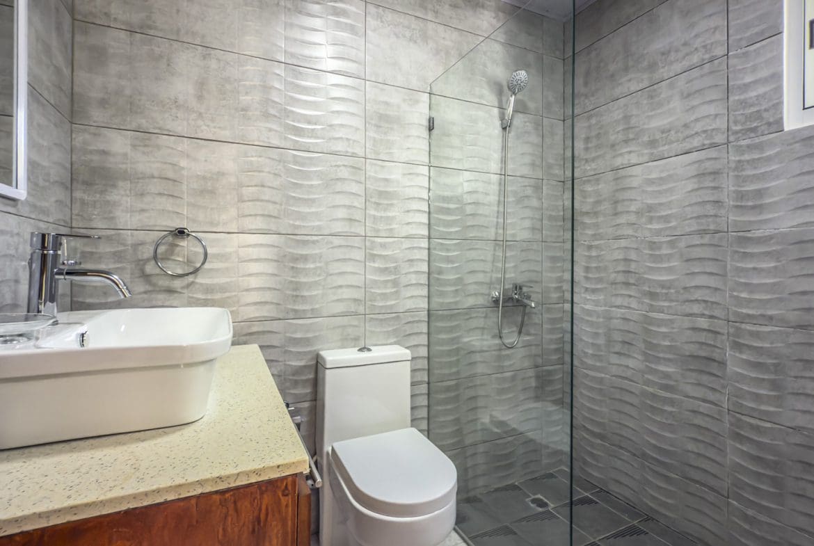 buyDRproperty Modern bathroom with textured gray tiles, white toilet, sink, and glass shower enclosure. Sosua Real Estate